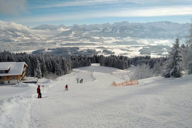 Vue du Mont Ultima. Ciel bleu, piste de ski enneigée avec un chalet et de la forêt, chaîne de montagnes au fond.